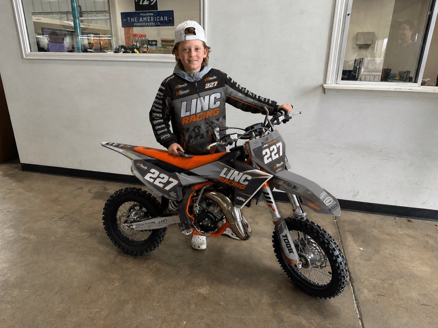 Lincoln Hauk standing with his number 227 LINC Racing dirt bike inside the shop.