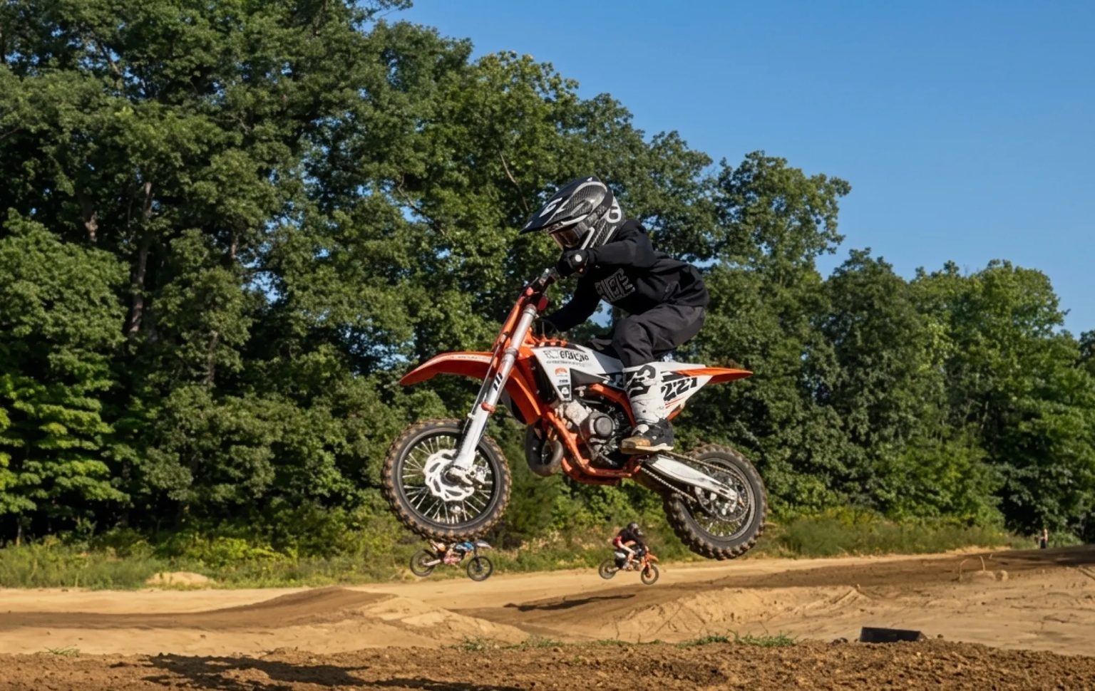 Lincoln Hauk, #227, airborne over a jump on his KTM at a motocross race, with young riders on the track behind him.